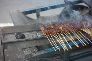 il satay venditore va in giro utilizzando un' carrello e fa sicuro il satay è grigliato propriamente. saziare è carne o pollo condito con bambù spiedini. foto