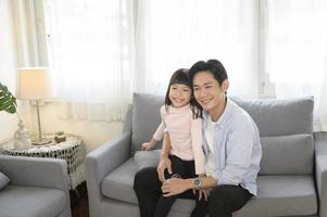 ritratto di asiatico padre e figlia seduta su divano nel il vivente camera foto