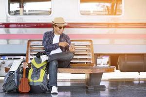 ritratto il turista giovane uomo viaggiatore con zaino sedersi in attesa treno stazione e pubblico auto Guarda a il lui mano uso il Telefono per dai un'occhiata il programma per partire viaggio a il stazione.viaggio concetto. foto