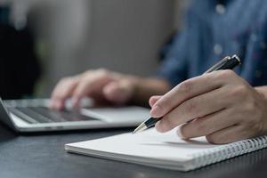 maschio mano scrittura nel taccuino con penna su il computer portatile computer nel moderno ufficio. foto