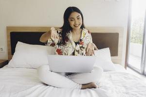 contento giovane donna Lavorando a partire dal il letto. hold il il computer portatile su il ginocchia e genere su il tastiera del computer. foto