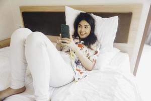 contento giovane donna controllo su sua Telefono mentre dire bugie nel letto foto