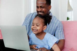 sorridente e contento africano padre indossare blu camicia con poco figlia utilizzando il computer portatile in linea shopping su divano. foto