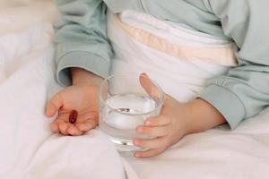 vicino su mani bambino nel letto detiene pillola e bicchiere di acqua assunzione medicinale, vitamina minerale integratori, alimentazione, salute cura, trattamento foto