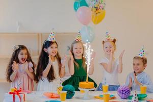 gli amici felicissimi dei bambini battono le mani e guardano la grande scintilla sulla torta, stanno vicino al tavolo festivo con regali, tazze e attributi delle vacanze, divertiti insieme, festeggia il compleanno foto