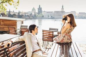 la bella fotografa professionista fa foto di suo marito che posa alla macchina fotografica sulla panca di legno al caffè all'aperto vicino alla bellissima vista panoramica della città antica. concetto di persone e hobby