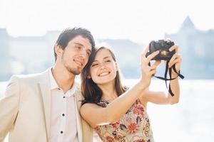 coppia felice innamorata posa per selfie nella moderna fotocamera professionale, cammina all'aperto, esplora luoghi sconosciuti nella grande città dei turisti, modella contro un paesaggio meraviglioso con acqua sullo sfondo foto
