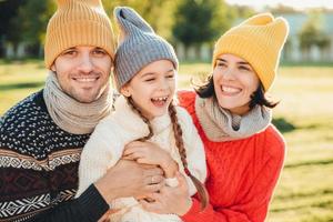 il bambino giocoso con le trecce indossa abiti caldi, trascorre il tempo libero con genitori adorabili e affettuosi, ha un'espressione felice, si sente rilassato. tre giovani membri della famiglia trascorrono piacevoli momenti foto