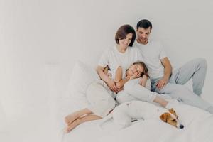 foto di una bella famiglia che trascorre il tempo libero insieme, sta a letto per molto tempo, ha un sonno salutare, il cane di razza giace vicino ai proprietari, gode di comfort e relax, spazio vuoto su sfondo chiaro per la promozione