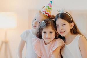 il ritratto di tre amici felici indossa abiti festivi, cappello da festa, posa al coperto su sfondo bianco, gioca insieme durante la festa di compleanno. le ragazze carine hanno un buon umore, vieni in un'occasione speciale foto