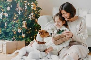 la giovane donna felice abbraccia la sua piccola figlia, tiene il regalo di Natale, attende le vacanze invernali, gioca con il cucciolo di razza, posa sul pavimento vicino all'albero di capodanno decorato. concetto di tempo di famiglia felice foto