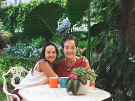 contento asiatico anziano donna e sua figlia seduta insieme a bianca tavolo nel bellissimo giardino, figlia abbracciare sua madre sorridente e guardare a telecamera. foto