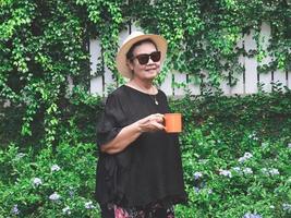salutare anziano asiatico donna indossare nero camicetta, occhiali da sole e cappello, Tenere arancia colore tazza di caffè nel il giardino con viola fiori, sorridente e guardare a telecamera. foto