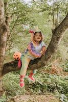 poco ragazza nel un' arcobaleno unicorno costume kigurumi è seduta su un' albero con zucca cestino per dolci contro il sfondo di un' foresta. Halloween concetto. spazio per testo. alto qualità foto
