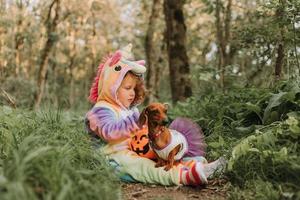 poco ridendo ragazza e un' nano bassotto nel Halloween costumi con un' zucca cestino per dolci all'aperto. un' ragazza nel un' arcobaleno unicorno kigurumi costume, un' cane nel un' vestito con un' pieno gonna. superiore Visualizza foto