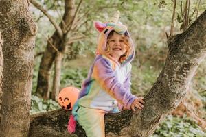 poco ragazza nel un' arcobaleno unicorno costume kigurumi è seduta su un' albero con zucca cestino per dolci contro il sfondo di un' foresta. Halloween concetto. spazio per testo. alto qualità foto