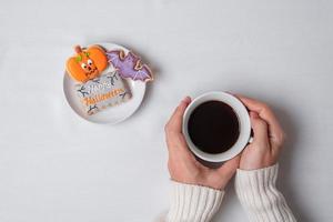 mano della donna che tiene la tazza di caffè durante il consumo di biscotti divertenti di halloween. buon giorno di halloween, trucco o minaccia, ciao ottobre, autunno autunno, tradizionale, festa e concetto di vacanza foto