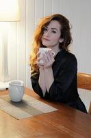 giovane donna nel condimento toga potabile tazza di caffè seduta a cucina tavolo foto