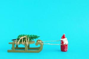 Santa Claus è trasporto un' Natale albero su un' verde di legno slitta su un' luminosa blu sfondo. Natale e nuovo anno concetto con copia spazio. foto