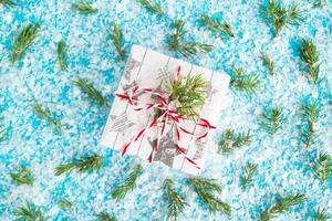 regalo scatola avvolto di bianca e grigio carta con rosso bianco nastro su il blu sfondo con artificiale neve decorato abete rami, superiore Visualizza. Natale e nuovo anno concetto con copia spazio. foto