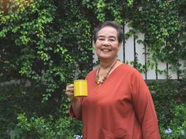 contento e salutare anziano asiatico donna Tenere giallo tazza di caffè in piedi nel il giardino, sorridente e guardare a telecamera. foto
