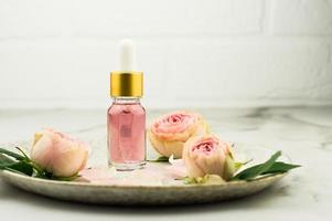un' bicchiere bottiglia con un' contagocce pipetta con essenziale olio di rosa petali su un' marmo tavolo di fronte un' bianca mattone parete. rosa mini cuffie su un' ceramica piatto. foto