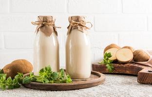 Due bottiglie di a base vegetale Patata latte su un' il giro di legno vassoio di fronte un' bianca mattone parete. tritato Patata tuberi su un' tavola. alternativa latte.