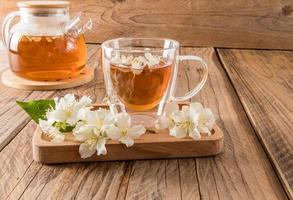 bicchiere trasparente tazza con fragrante gelsomino tè su un' di legno vassoio con il fiori di il pianta. di legno rustico tavolo con teiera. tè composizione. foto