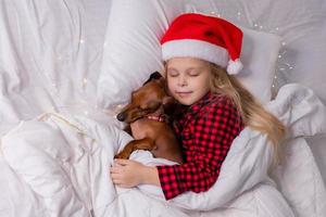 poco ragazza dorme nel letto con un' bassotto cane per Natale foto