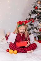 contento poco bionda ragazza a casa nel pigiama per Natale ordinamento regali e giocando con Natale giocattoli foto