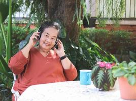 contento e salutare asiatico anziano donna seduta con caffè tazza e fiore pentole nel all'aperto giardino, ascoltando per preferito musica a partire dal cuffia, sorridente e guardare a telecamera. foto