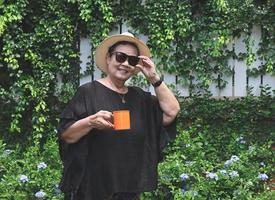 salutare asiatico anziano donna indossare nero camicetta, occhiali da sole e cappello, Tenere arancia colore tazza di caffè, in piedi nel il giardino con viola fiori, , sorridente e guardare a telecamera. foto