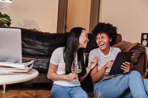 divertente donne nel jeans avere divertimento, bevanda Champagne e Guarda a tavoletta mentre seduta nel appartamento. foto