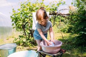 la bambina in età prescolare aiuta con il bucato. il bambino lava i vestiti in giardino foto