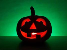 Halloween lavorato raggiante zucca jack-o-lanterna con candela dentro sta su superficie nel buio notte e sfocato sfocato sfondo. il indietro parete evidenziato nel verde. spaventoso vacanza concetto. foto