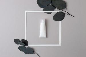 bianca tubo di cosmetico crema con bianca telaio e eucalipto su grigio sfondo. piatto posare foto
