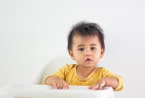 bambino ragazza di asiatico etnia giallo camicia si siede pazientemente nel un' sedia solo a casa. espressione di emozione, eccitazione, infanzia, e infantilismo. su quello carino Wow viso felicità vita foto