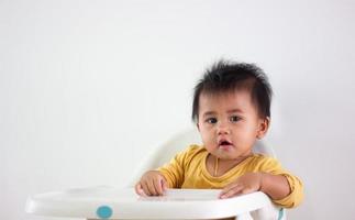 bambino ragazza di asiatico etnia giallo camicia si siede pazientemente nel un' sedia solo a casa. espressione di emozione, eccitazione, infanzia, e infantilismo. su quello carino Wow viso felicità vita foto