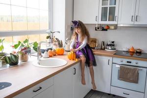 bambino decora il cucina nel casa per Halloween. ragazza nel un' strega costume giochi con il arredamento per il vacanza - pipistrelli, Jack lanterna, zucche. autunno comfort nel Casa, in stile scandinavo cucina, soffitta foto