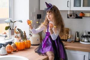 bambino decora il cucina nel casa per Halloween. ragazza nel un' strega costume giochi con il arredamento per il vacanza - pipistrelli, Jack lanterna, zucche. autunno comfort nel Casa, in stile scandinavo cucina, soffitta foto