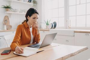 femmina casalinga libero professionista lavori a computer portatile, piani acquisti, Appunti, seduta nel cucina a casa foto