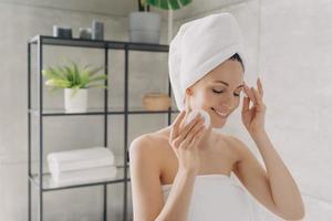 pelle purificazione, cura della pelle. sorridente femmina pulizia facciale pelle di cotone pastiglie nel bagno dopo doccia foto