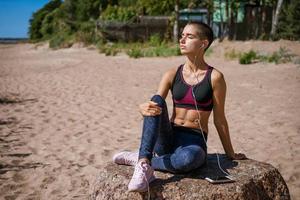 giovane donna vestito nel abbigliamento sportivo si siede su spiaggia per apprendimento musica ascoltando foto