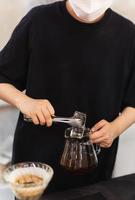 barista con viso maschera fabbricazione ghiacciato caffè di mettendo un ghiaccio in un' bicchiere vaso con nero appena fermentato caffè. foto