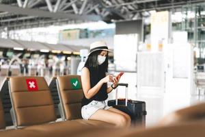 adulto turista donna con bagaglio indossare maschera per covid-19 dai un'occhiata nel con mobile Telefono a aeroporto terminale. foto