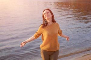 contento donna rilassa con sua braccia su per lati su riva lago a tramonto nel raggi foto