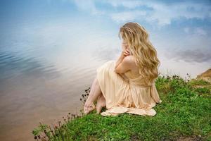 bellissimo triste ragazza seduta su il riva di il lago nel un' vestito , il concetto di solitudine foto