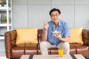 anziano asiatico uomini seduta su il divano a casa mentre mangiare verde verdura insalata e biologico arancia succo per prima colazione. salutare mangiare stile di vita per Sambuco la pensione persone concetto. foto