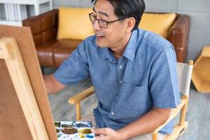 interno tiro di anziano asiatico uomo sedersi e pittura immagine su cavalletto dentro di il Casa. tempo libero attività per anziano per rilassamento, contento la pensione stile di vita concetto. foto