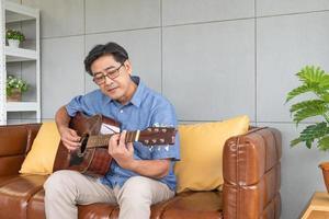 asiatico Sambuco uomo godere giocando chitarra a il divano dentro di bene interno decorazione Casa. attivo anziano stile di vita dopo la pensione con felicità con un' bene mentale Salute. foto